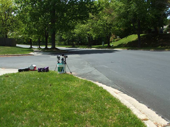 We are looking to the left along a two-lane street with curbs, grass and trees on both sides.  The road is going slightly downhill and curves gently to the left, disappearing about 100 feet away.  About 20 feet from us is the corner of another street.  A tripod about 2 feet high and some equipment lie on the grass.